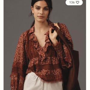 May include: A brown and red floral print blouse with a ruffled V-neck and long sleeves. The blouse has a semi-sheer look and is worn with white trousers. A brown leather tote bag is carried over the shoulder.