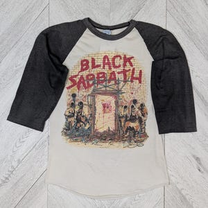 May include: A white and gray baseball tee shirt with a black and red graphic of the band Black Sabbath. The graphic features a brick wall with a gate and figures in front of it.