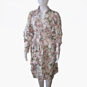 May include: A cream-coloured dress with a floral pattern. The dress has long sleeves, a collared neckline, and a matching belt. The floral design includes red, green, and brown elements. The dress is buttoned down the front and has a knee-length hemline.