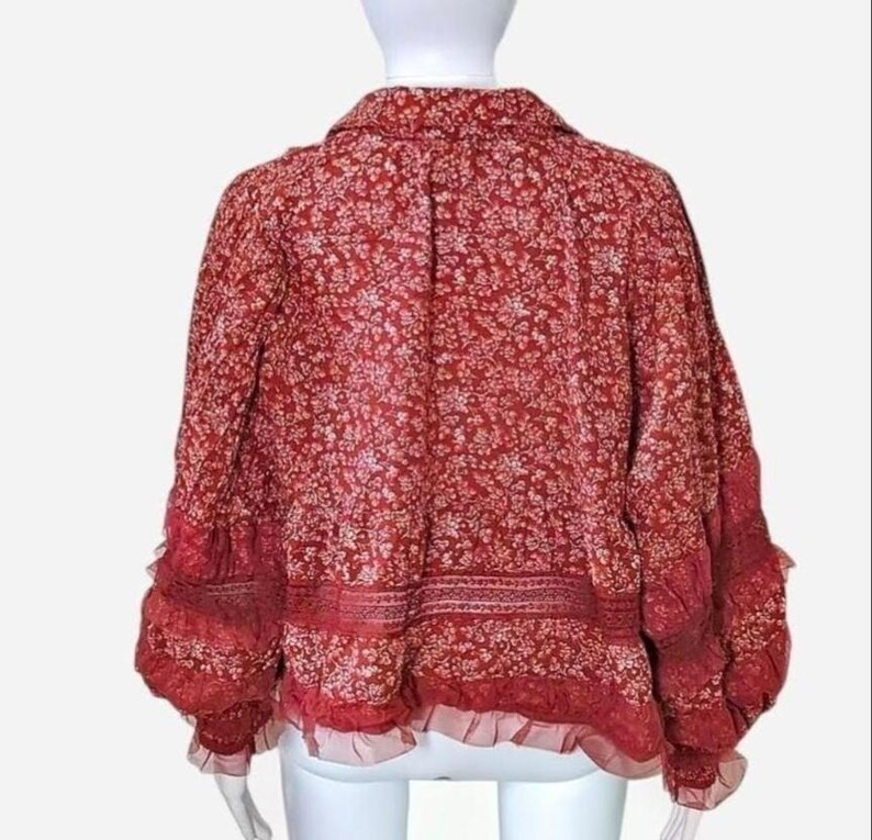 May include: A red floral print blouse with a collared neckline and long sleeves. The blouse has a ruffled hem and cuffs, and a delicate, all-over white floral pattern. The fabric appears lightweight and flowy, suitable for warmer weather.