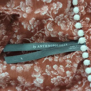 May include: Close-up of a rust-coloured fabric with a white floral pattern. The tag reads "by ANTHROPOLOGIE" and "made in ind (fabrique en)". White pom-poms line the edge of the fabric. The size is L.