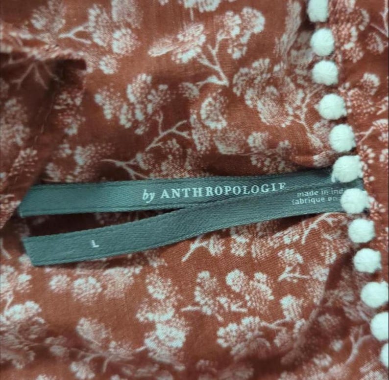 May include: Close-up of a rust-coloured fabric with a white floral pattern. The tag reads "by ANTHROPOLOGIE" and "made in ind (fabrique en)". White pom-poms line the edge of the fabric. The size is L.