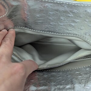 Puede incluir: Bolso de mano plateado con un patr&oacute;n texturizado tipo avestruz. El interior es gris claro y el bolso tiene cierre de cremallera. El bolso est&aacute; abierto, revelando el interior.
