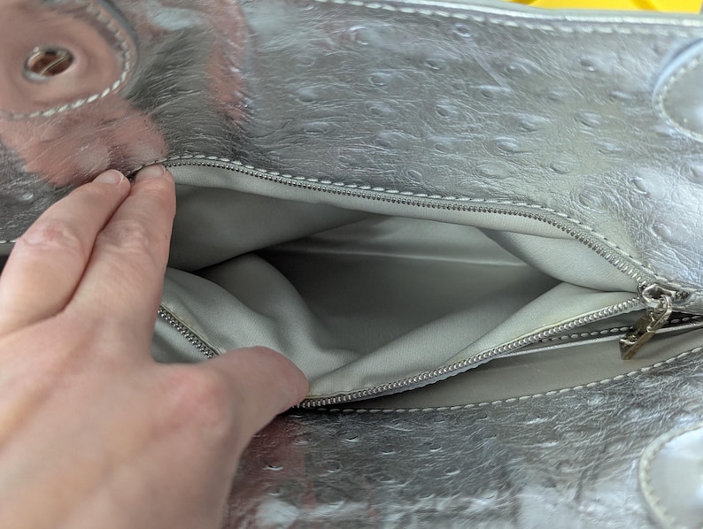 Puede incluir: Bolso de mano plateado con un patr&oacute;n texturizado tipo avestruz. El interior es gris claro y el bolso tiene cierre de cremallera. El bolso est&aacute; abierto, revelando el interior.
