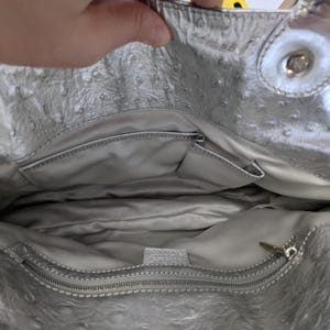 Puede incluir: Bolso de mano plateado con exterior texturizado y cierre de cremallera. El interior cuenta con m&uacute;ltiples bolsillos y forro plateado. El bolso tiene un asa plateada y un cierre a presi&oacute;n.
