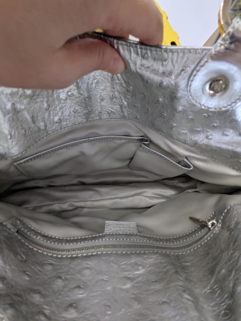 Puede incluir: Bolso de mano plateado con exterior texturizado y cierre de cremallera. El interior cuenta con m&uacute;ltiples bolsillos y forro plateado. El bolso tiene un asa plateada y un cierre a presi&oacute;n.