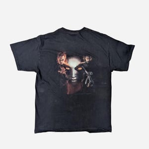 May include: Black t-shirt featuring a graphic design of a face with glowing orange eyes and two figures on either side. The design has a dark, ominous theme. The shirt is made of a soft, dark fabric.