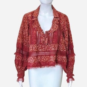 May include: A red floral print blouse with long sleeves and a V-neck. The blouse has a ruffled collar and cuffs, and a lace panel across the chest. The fabric has a delicate, semi-sheer appearance.