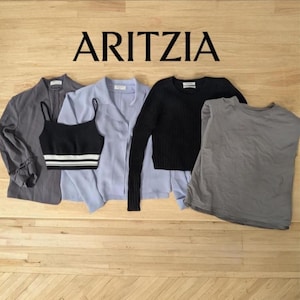 Può includere: Una collezione di capi d'abbigliamento femminile, tra cui un blazer grigio, un top corto nero con strisce bianche, una camicia azzurra con bottoni, un maglione nero a maniche lunghe e una canotta grigia. Il marchio "ARITZIA" è visualizzato in alto.