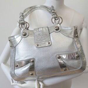 Puede incluir: Bolso de mano plateado con un acabado met&aacute;lico brillante. El bolso presenta un dise&ntilde;o estructurado con un asa superior, herrajes plateados y remaches decorativos. El bolso tiene forma rectangular con cierre de solapa.