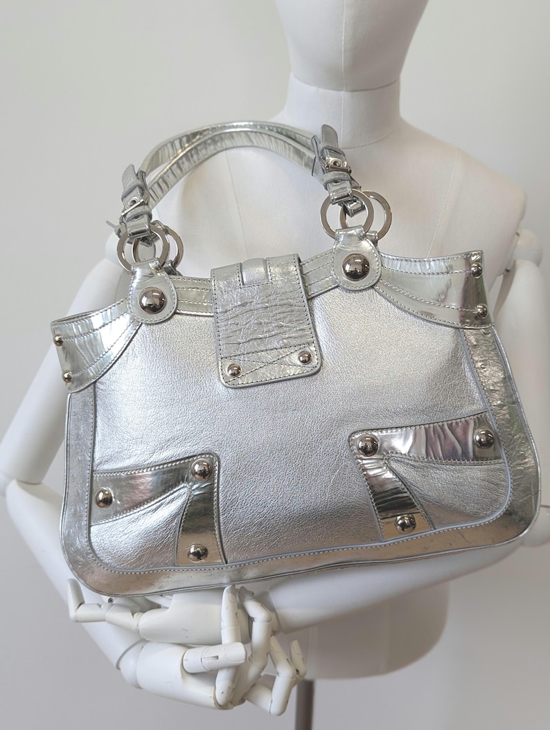 Puede incluir: Bolso de mano plateado con un acabado met&aacute;lico brillante. El bolso presenta un dise&ntilde;o estructurado con un asa superior, herrajes plateados y remaches decorativos. El bolso tiene forma rectangular con cierre de solapa.