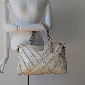 Puede incluir: Bolso Coach con estampado de piel de serpiente en tonos crema y gris. El bolso presenta un diseño acolchado, asas superiores dobles y herrajes plateados. Una pequeña etiqueta cuelga del asa. El bolso se exhibe en un maniquí.
