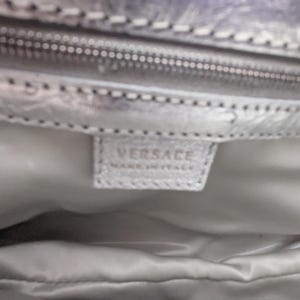 Puede incluir: Primer plano del interior de un bolso plateado. La imagen muestra una cremallera, un forro de tela y una peque&ntilde;a etiqueta rectangular con la marca "VERSACE" y "MADE IN ITALY" impresa.