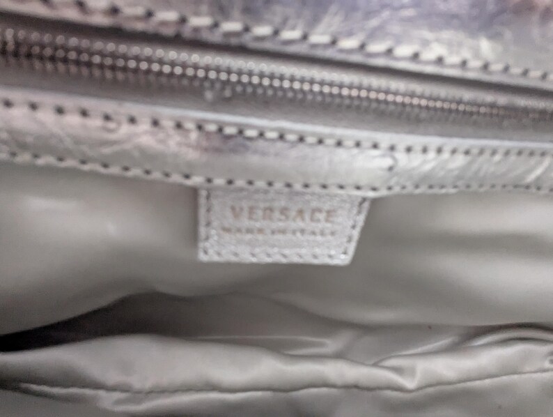 Puede incluir: Primer plano del interior de un bolso plateado. La imagen muestra una cremallera, un forro de tela y una peque&ntilde;a etiqueta rectangular con la marca "VERSACE" y "MADE IN ITALY" impresa.