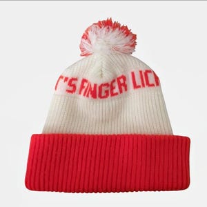 Pode incluir: Um gorro branco de malha com uma aba vermelha e um pompom vermelho e branco. O texto "IT'S FINGER LICK" está impresso em vermelho na frente do chapéu. O chapéu é feito de um material de malha macia.