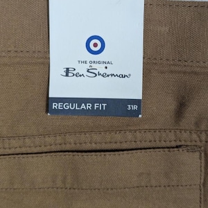 May include: Close-up of brown trousers with a tag. The tag reads "THE ORIGINAL Ben Sherman" with a bullseye logo. The tag also says "REGULAR FIT" and "31R".