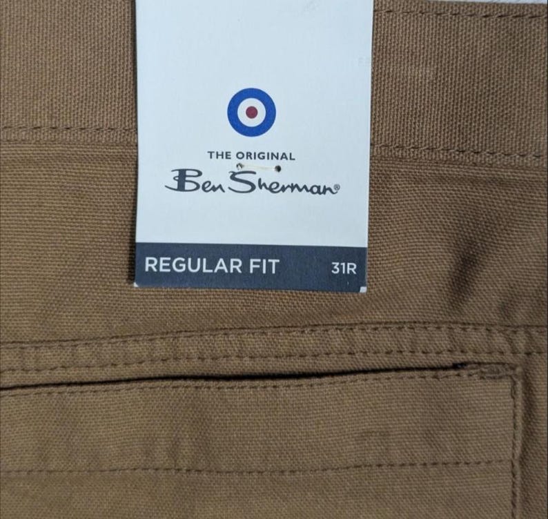 May include: Close-up of brown trousers with a tag. The tag reads "THE ORIGINAL Ben Sherman" with a bullseye logo. The tag also says "REGULAR FIT" and "31R".