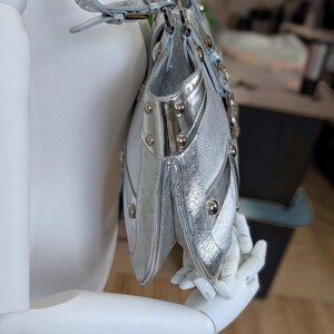 Puede incluir: Bolso plateado con acabado texturizado, con m&uacute;ltiples compartimentos y tachuelas met&aacute;licas decorativas. El bolso tiene una correa para el hombro y se exhibe en un maniqu&iacute;. El dise&ntilde;o del bolso incluye detalles plateados y una forma estructurada.