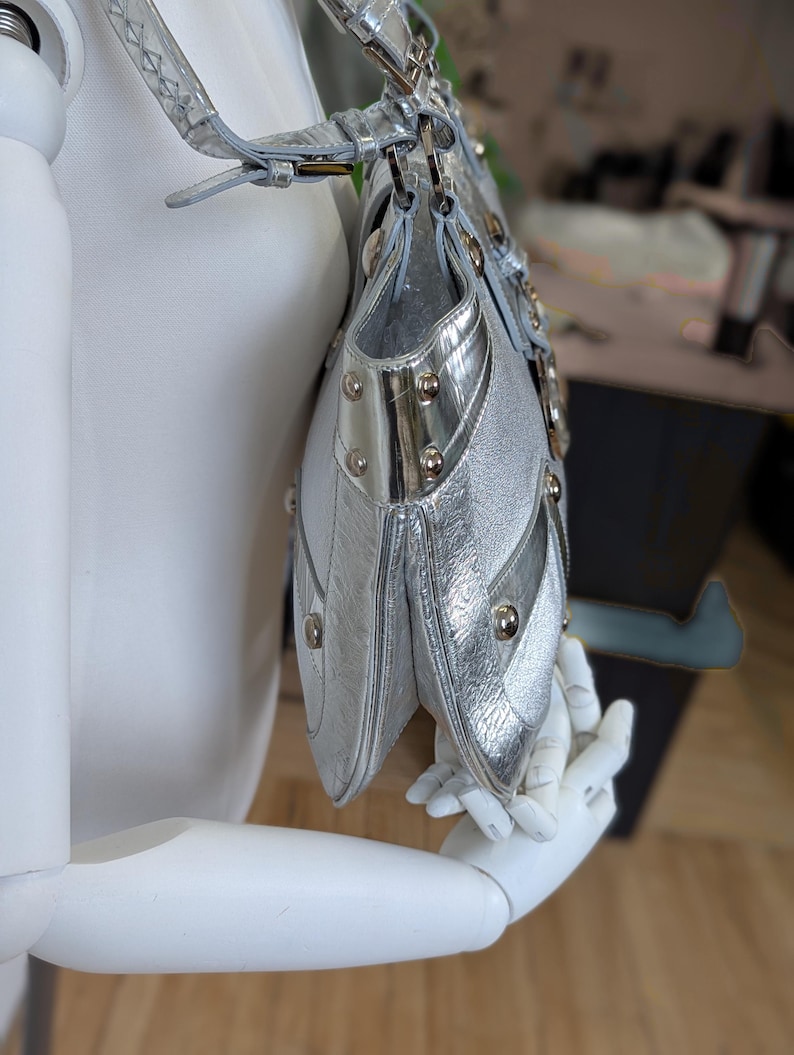 Puede incluir: Bolso plateado con acabado texturizado, con m&uacute;ltiples compartimentos y tachuelas met&aacute;licas decorativas. El bolso tiene una correa para el hombro y se exhibe en un maniqu&iacute;. El dise&ntilde;o del bolso incluye detalles plateados y una forma estructurada.