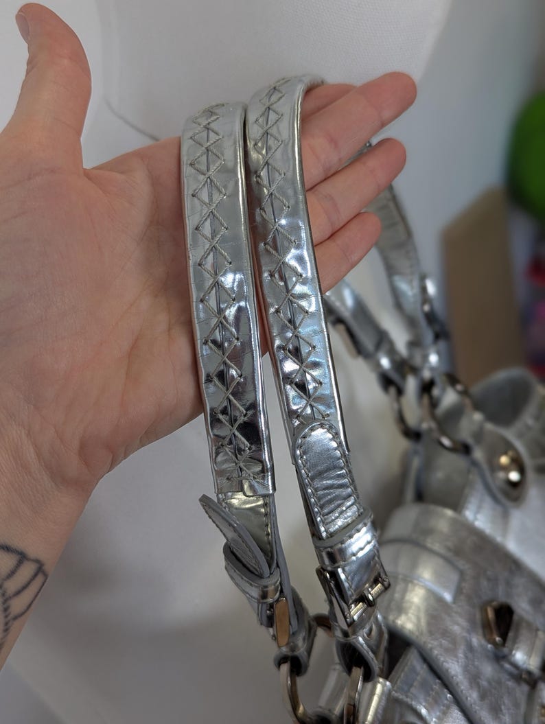 Puede incluir: Bolso de mano plateado con asas trenzadas. Las asas son de material met&aacute;lico brillante con un patr&oacute;n de diamantes. El bolso tiene un acabado plateado y herrajes met&aacute;licos.