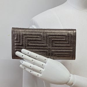 May include: A metallic bronze wallet with a geometric pattern. The wallet is rectangular and features a repeating, stitched design. The color is a muted bronze with a slight sheen. The wallet is held by a white mannequin hand.