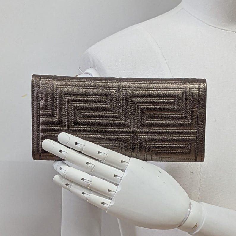 May include: A metallic bronze wallet with a geometric pattern. The wallet is rectangular and features a repeating, stitched design. The color is a muted bronze with a slight sheen. The wallet is held by a white mannequin hand.