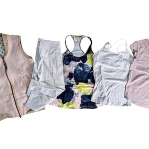 May include: A collection of women's athletic wear, including a light pink zip-up vest, white leggings, a patterned tank top with blue and yellow accents, a white camisole, and a light pink V-neck shirt. The items are arranged on a white background.