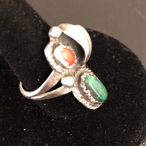 Op de afbeelding: Een zilveren ring met een groene en rode stenen setting. De ring ligt op een zwarte fluwelen achtergrond.