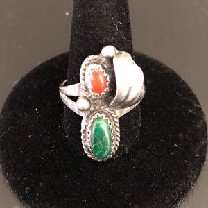 Op de afbeelding: Een zilveren ring met een groene en rode stenen setting. De ring heeft een uniek ontwerp met een gebogen zilveren band en een decoratieve swirl.