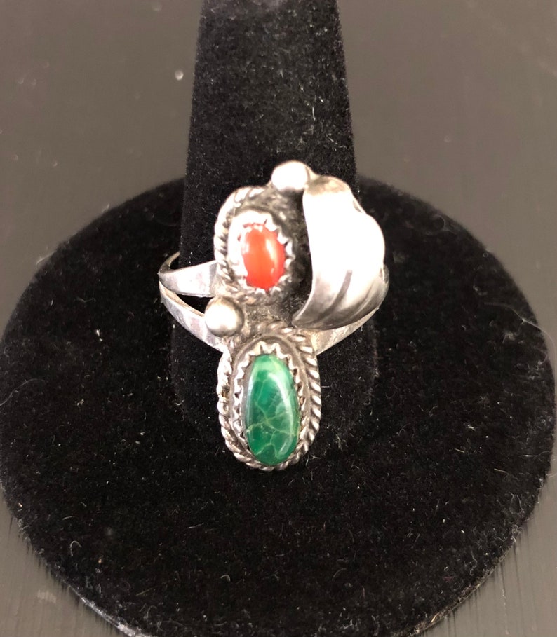 Op de afbeelding: Een zilveren ring met een groene en rode stenen setting. De ring heeft een uniek ontwerp met een gebogen zilveren band en een decoratieve swirl.