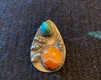 May include: A silver pendant with a teardrop shape features a carved design of leaves and two stones. The top stone is a turquoise blue and the bottom stone is a banded agate with orange and white stripes.