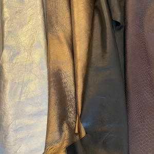 Könnte beinhalten: Nahaufnahme von mehreren Stoffrollen in verschiedenen Farben. Die Stoffe umfassen metallisches Silber, Gold, Dunkelbronze und einen strukturierten Stoff mit Schlangenmuster in einem gedämpften Lila. Die Stoffe scheinen Leder oder Kunstleder zu sein.