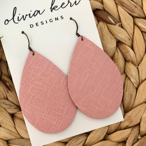 May include: A pair of pink teardrop-shaped earrings with a textured surface. The earrings are made of leather and have a silver hook closure. The earrings are displayed on a white card with the text "olivia keri DESIGNS".