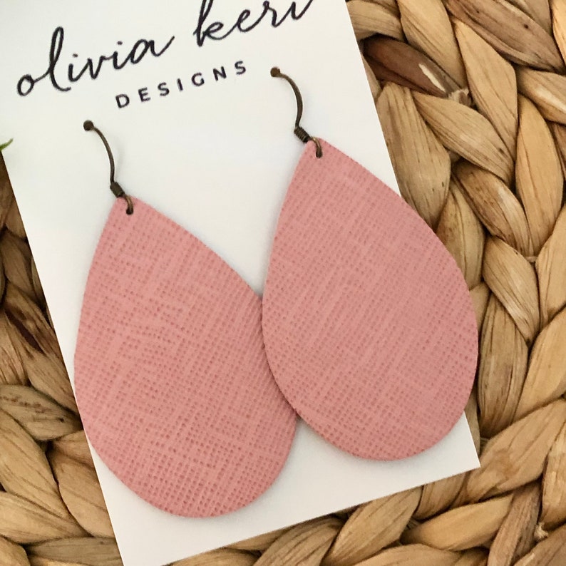 May include: A pair of pink teardrop-shaped earrings with a textured surface. The earrings are made of leather and have a silver hook closure. The earrings are displayed on a white card with the text "olivia keri DESIGNS".