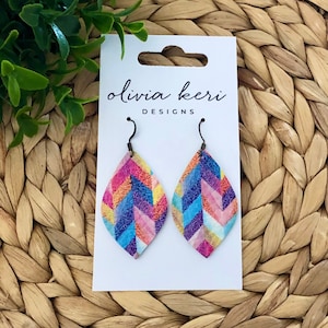 May include: A pair of colorful teardrop-shaped earrings with a chevron pattern. The earrings are made of leather and have silver hooks. The earrings are on a white card with the text "olivia keri DESIGNS".