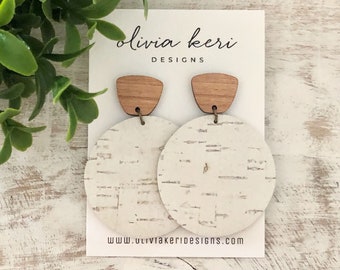 May include: A pair of earrings with a natural, minimalist design. Each earring features a light brown wooden top and a large, round, off-white cork base. The card behind the earrings reads "olivia keri DESIGNS" and "www.oliviakeridesigns.com".