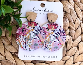 May include: A pair of floral print earrings with wooden backs. The earrings are round and feature a white background with pink, red, and yellow flowers. The text "olivia keri DESIGNS" and "www.oliviakeridesigns.com" are printed on the card behind the earrings.