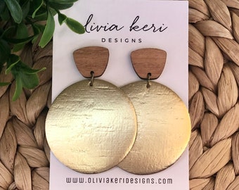 May include: Gold and wood earrings with a textured gold finish. The earrings are round and have a wooden top. The earrings are displayed on a white card with the text "livia keri DESIGNS" and "www.oliviakeridesigns.com".