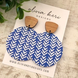 May include: A pair of dangle earrings featuring a wooden trapezoidal top piece connected to a large circular blue and white chevron patterned disc. The earrings are displayed on a white card labeled olivia keri DESIGNS www.oliviakeridesigns.com.