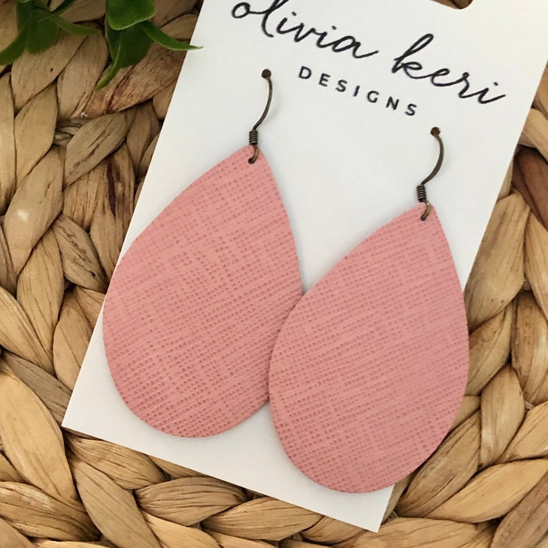 May include: A pair of pink teardrop-shaped leather earrings with a textured surface. The earrings are hanging from a white card with the text "Olivia Keri Designs".