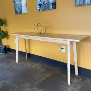 May include: A light-colored wooden console table with a rectangular top and four angled legs. A gold-colored abstract sculpture sits on the table. The table is approximately 72 inches long.