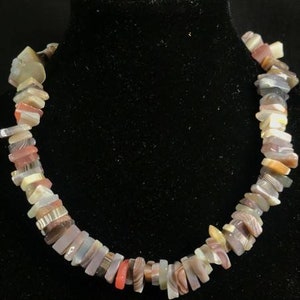 Puede incluir: Un collar hecho de cuentas de ágata multicolores. Las cuentas son rectangulares y tienen un patrón de rayas en capas. El collar está sobre un fondo negro.
