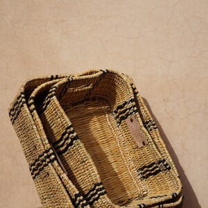 May include: Three rectangular woven baskets stacked together. The baskets are made of natural-colored straw with black stripes. A small leather tag is attached to the top basket. The baskets are on a woven chair.