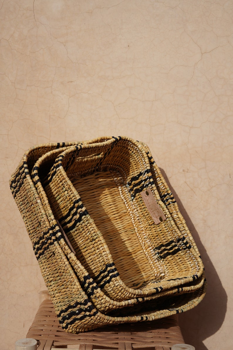 May include: Three rectangular woven baskets stacked together. The baskets are made of natural-colored straw with black stripes. A small leather tag is attached to the top basket. The baskets are on a woven chair.