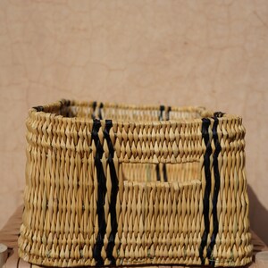 May include: A rectangular woven basket with a natural beige color, accented by vertical black stripes. The basket has a rectangular opening and is placed on a wooden surface.