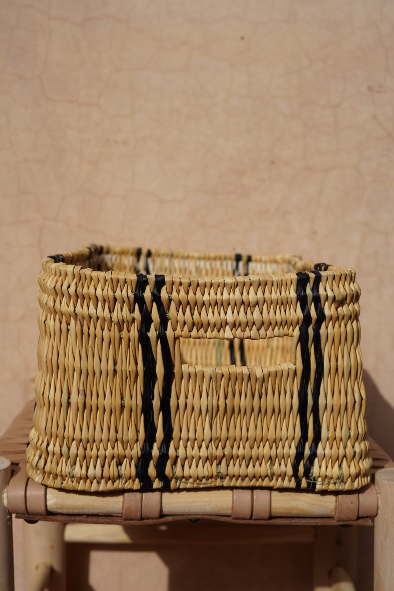 May include: A rectangular woven basket with a natural beige color, accented by vertical black stripes. The basket has a rectangular opening and is placed on a wooden surface.