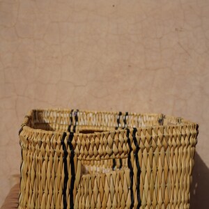 May include: A rectangular woven basket with a natural, light brown color. Vertical black stripes are woven into the sides. The basket has two rectangular cut-out handles. The basket is sitting on a wooden surface.