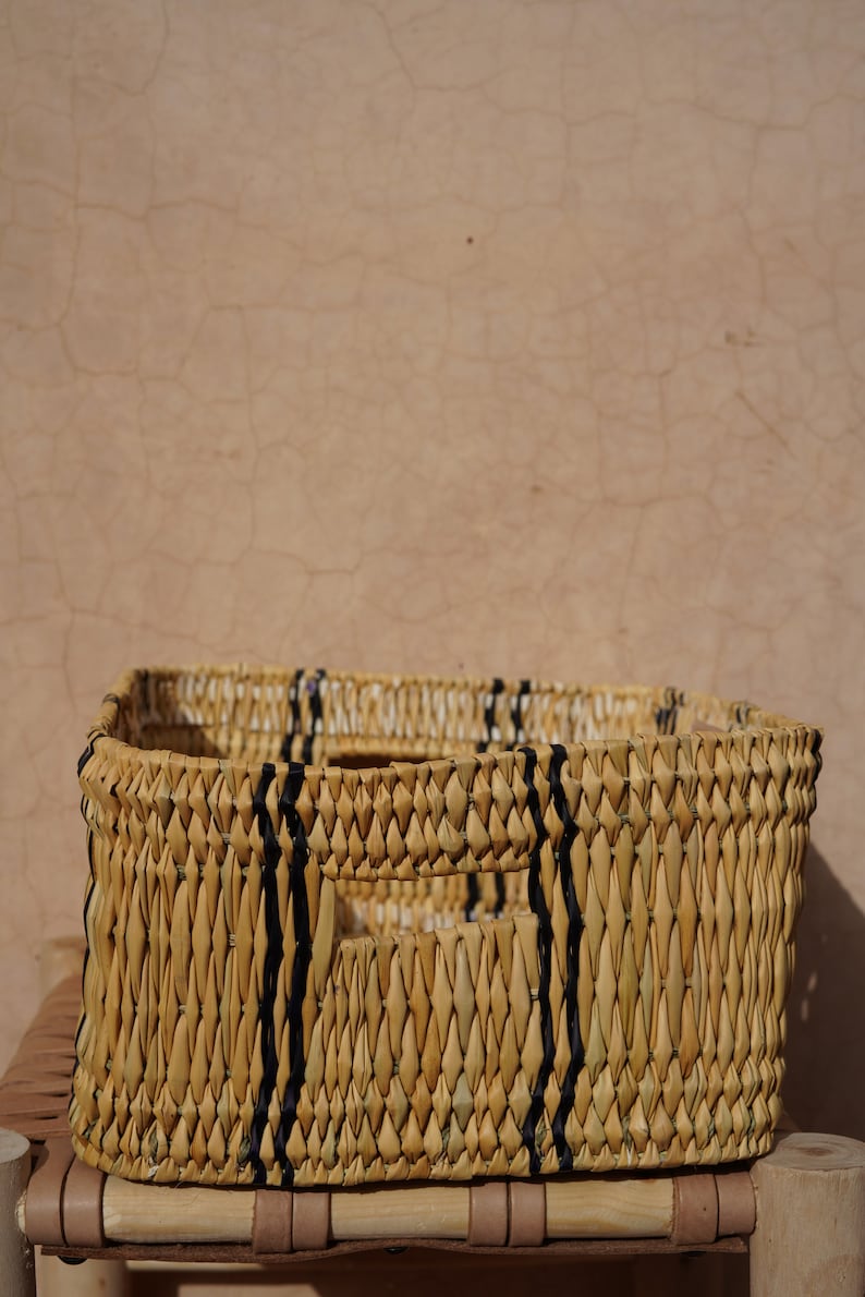 May include: A rectangular woven basket with a natural, light brown color. Vertical black stripes are woven into the sides. The basket has two rectangular cut-out handles. The basket is sitting on a wooden surface.
