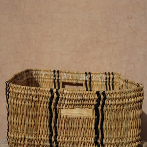 May include: A rectangular woven basket with a natural tan color and black accents. The basket has a square shape with handles and is sitting on a wooden stool. The background is a textured, light brown wall.