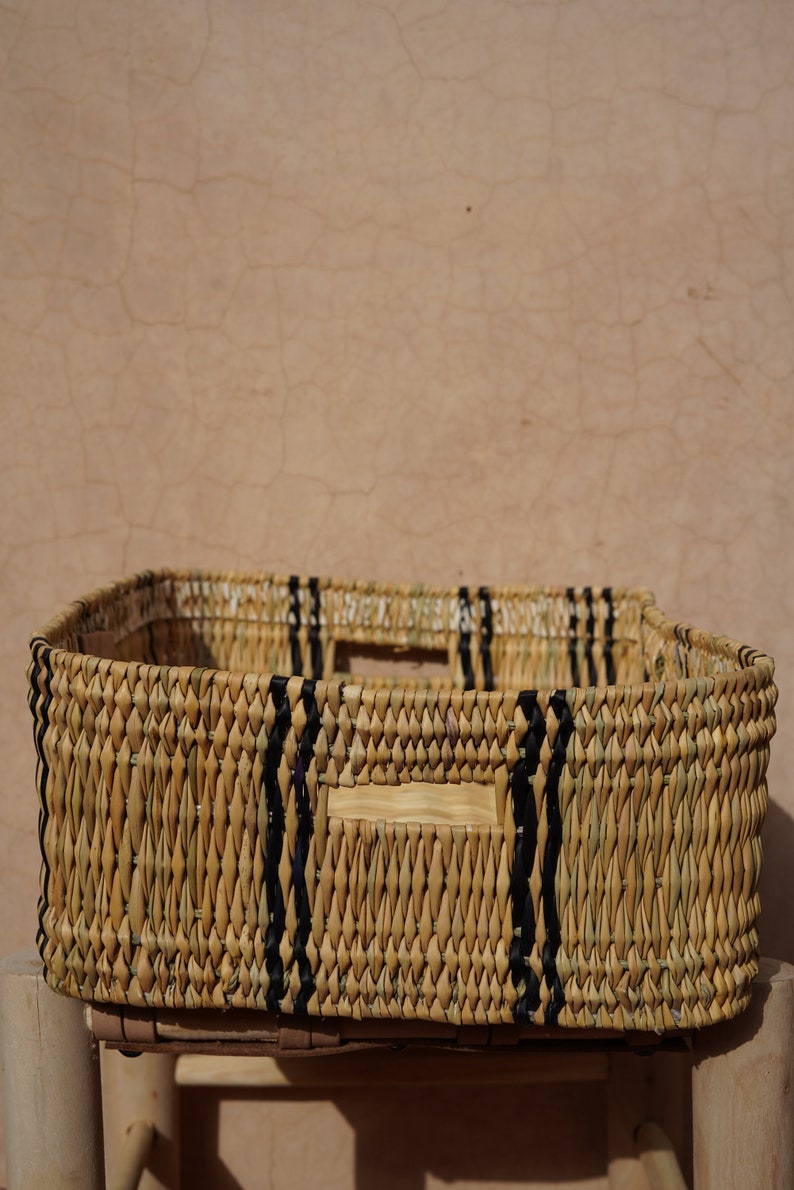 May include: A rectangular woven basket with a natural tan color and black accents. The basket has a square shape with handles and is sitting on a wooden stool. The background is a textured, light brown wall.